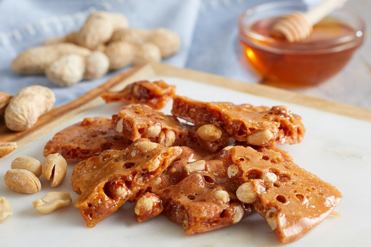 Caramel peanut brittle with peanuts and nuts, served on a white marble board with peanuts, a blue cloth, and a small bowl of honey in the background.