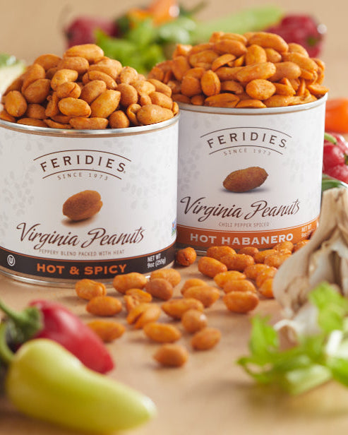 Two cans of Feridies Virginia Peanuts Hot & Spicy and Hot Habanero peanuts are displayed on a wooden table surrounded by peanuts, garlic, celery, and a blurred drink in the foreground.