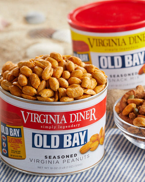 Two cans of Old Bay Seasoned Virginia Peanuts with a small glass bowl of peanuts, styled on a striped napkin on a light wooden surface.