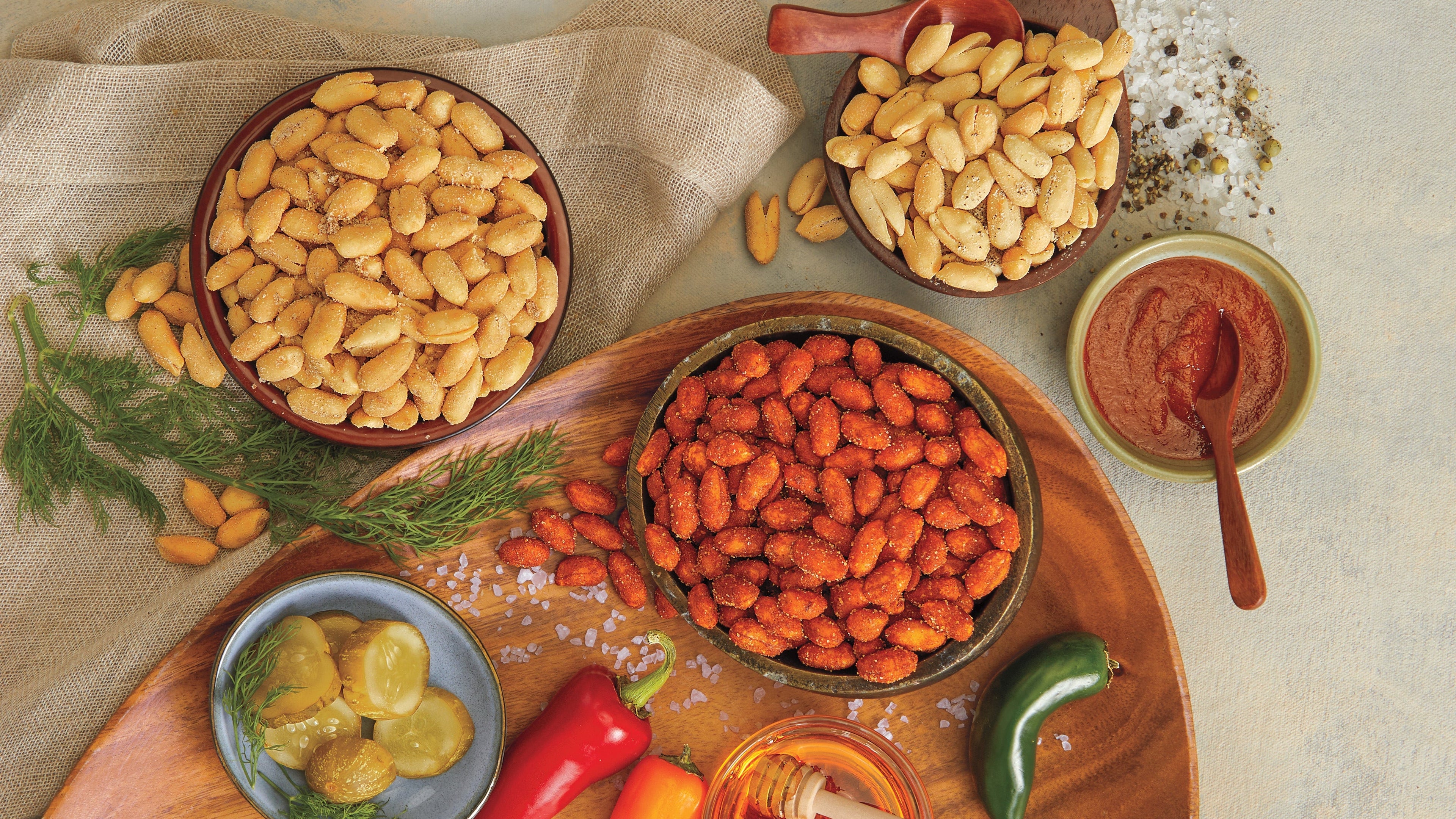 An overhead styled food scene on a light stone surface showing bowls of peanuts, red chili peppers, pickled jalapeños, a small bowl of red sauce, a honey dipper, and whole spices arranged on a wooden board with a napkin.